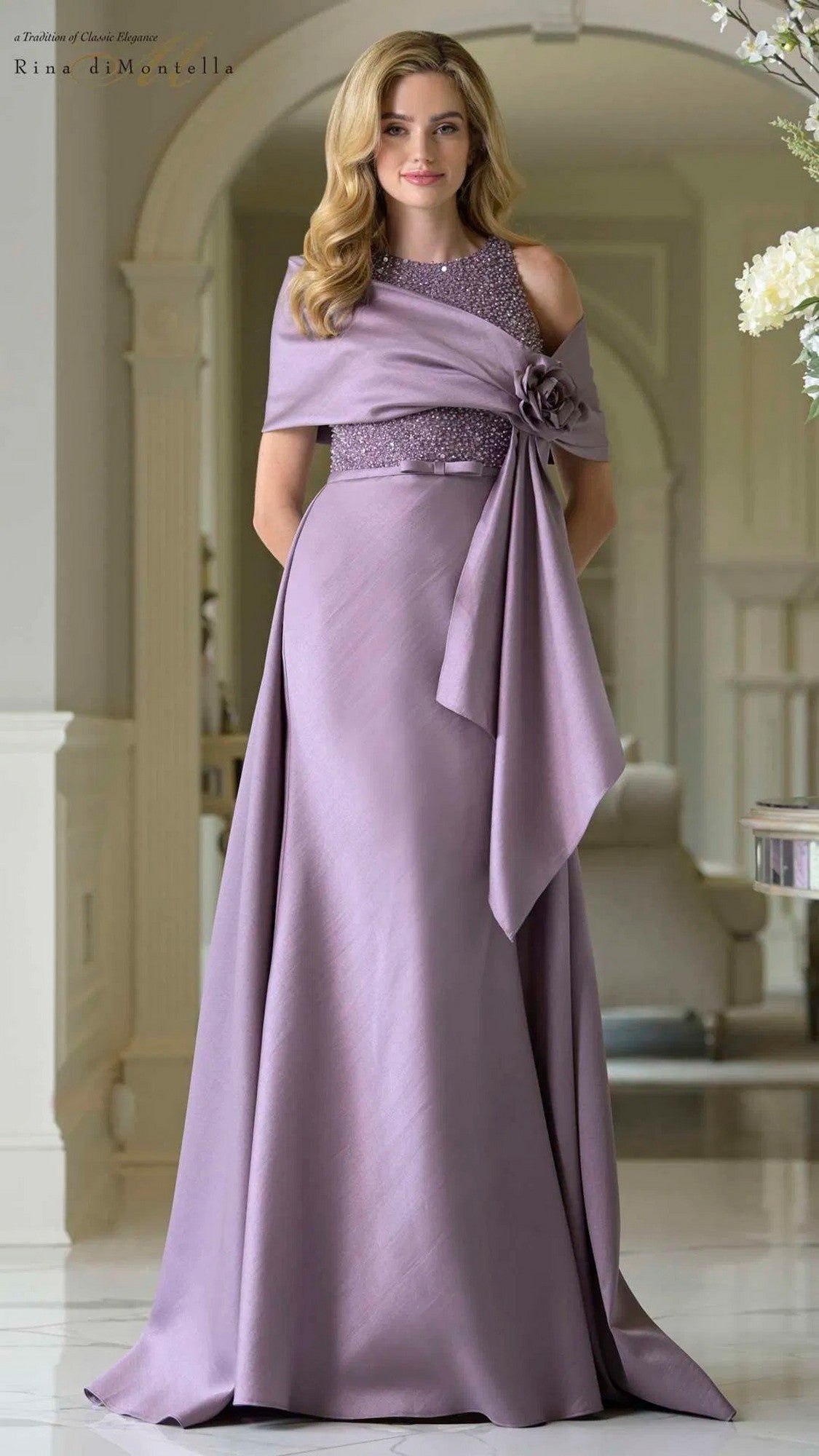 A woman stands indoors wearing the Rina Di Montella RD3404 Long Formal Dress in Victorian Lilac, featuring a beaded bodice, floral waist detail, and a draped shawl.